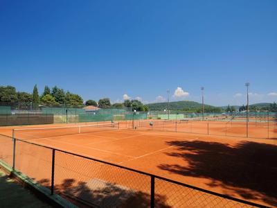 Slunečný tenisový kurt s červenou hlínou a oplocením, obklopený stromy a kopci.