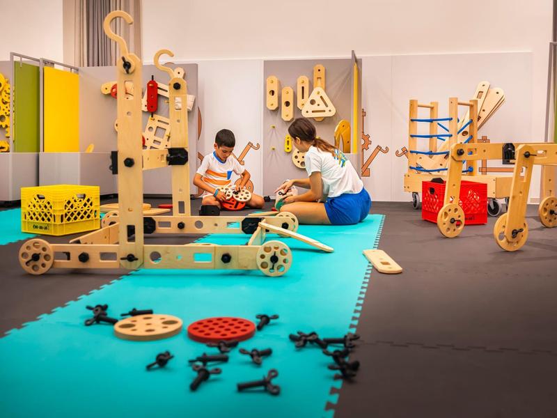 Des enfants jouent avec des jouets en bois dans une pièce lumineuse et colorée avec des tapis doux.