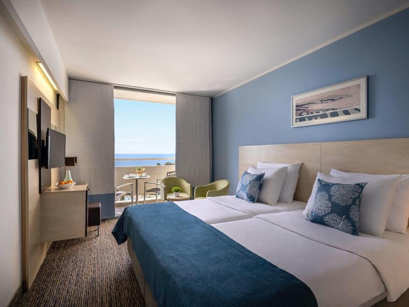 Chambre d'hôtel moderne avec vue sur la mer, lit double, mur bleu et coin salon près de la fenêtre.