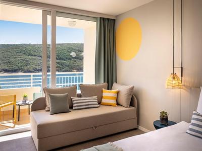Hotelkamer met bed, geel-grijze bank, balkon met uitzicht op zee en groen.