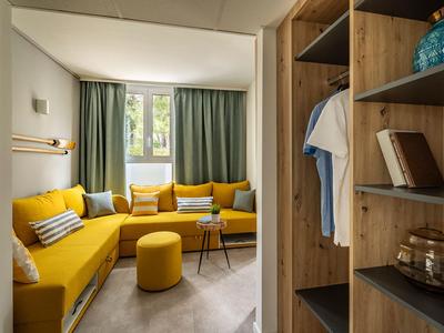 Gezellige zithoek met gele bank, kussens en houten meubels in een lichte hotelkamer.