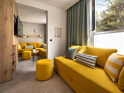 Kleine hotelkamer met gele bank, poefjes en zitgedeelte op de achtergrond.