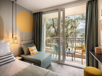 Lichte hotelkamer met tweepersoonsbed, fauteuil, balkon en uitzicht op bomen.