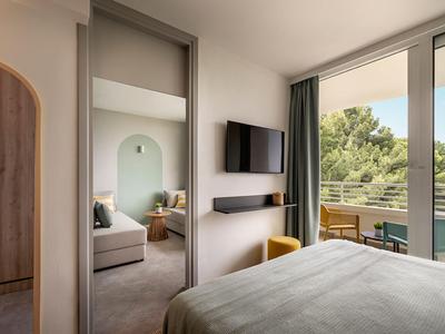 Modern hotelkamer met groot bed, spiegel, tv en balkon met uitzicht op bomen.