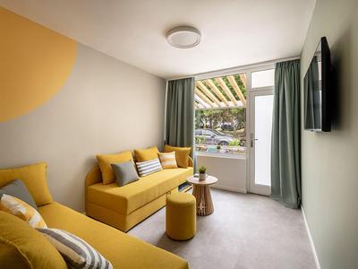 Licht ingericht hotelkamer met gele banken en toegang tot balkon.