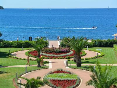 Blick auf gepflegte Gartenanlagen mit Blumenbeeten und Palmen vor dem blauen Meer.