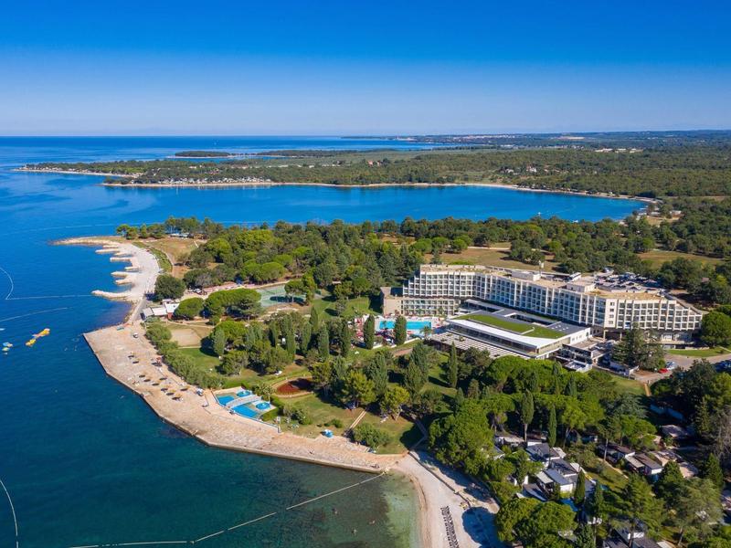 Aerial view of a hotel with pool on a forested coastline and sandy beach by the sea.