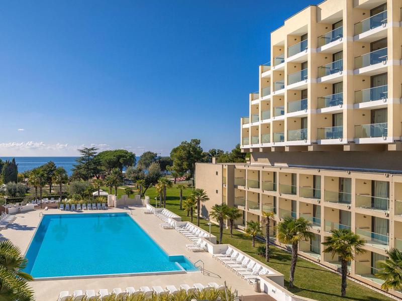 Hotel with large outdoor pool, sun terrace, and sea view under clear blue sky.
