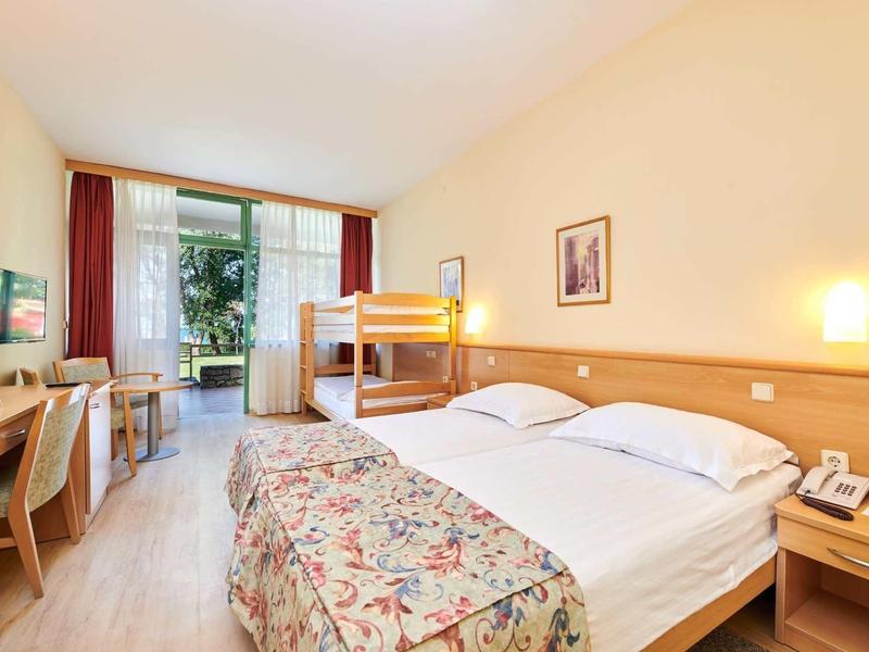 Chambre d'hôtel lumineuse avec lit double, lit superposé, bureau et balcon avec vue sur la verdure.