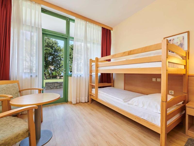 Chambre d'hôtel lumineuse avec lits superposés, coin salon et grande fenêtre donnant sur la verdure.