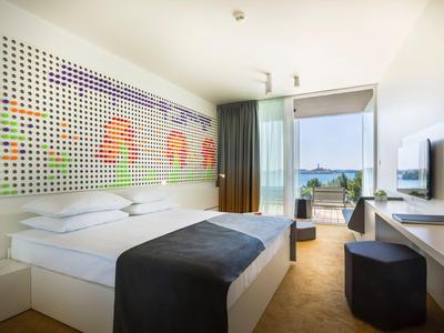Modernes Hotelzimmer mit Doppelbett, farbenfrohem Wandbild und Balkon mit Blick aufs Wasser.
