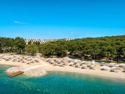 Beach with sun loungers and umbrellas by clear blue water and hotel at forest edge.