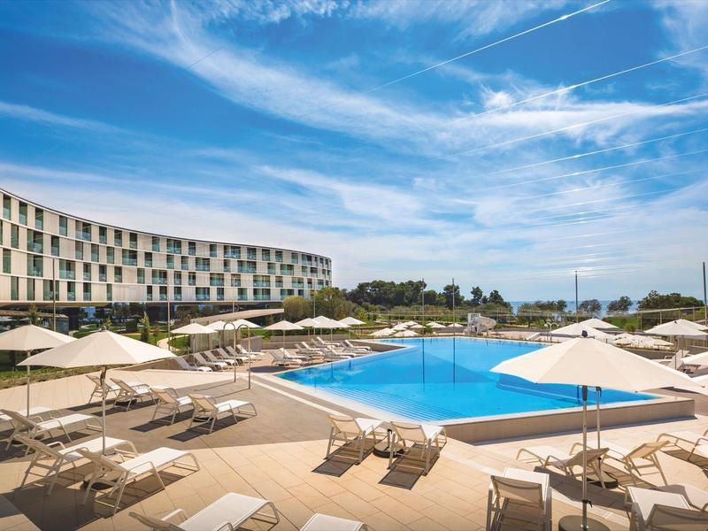 Moderner Außenpool mit Liegestühlen und weißen Sonnenschirmen vor einem gebogenen Hotelgebäude.