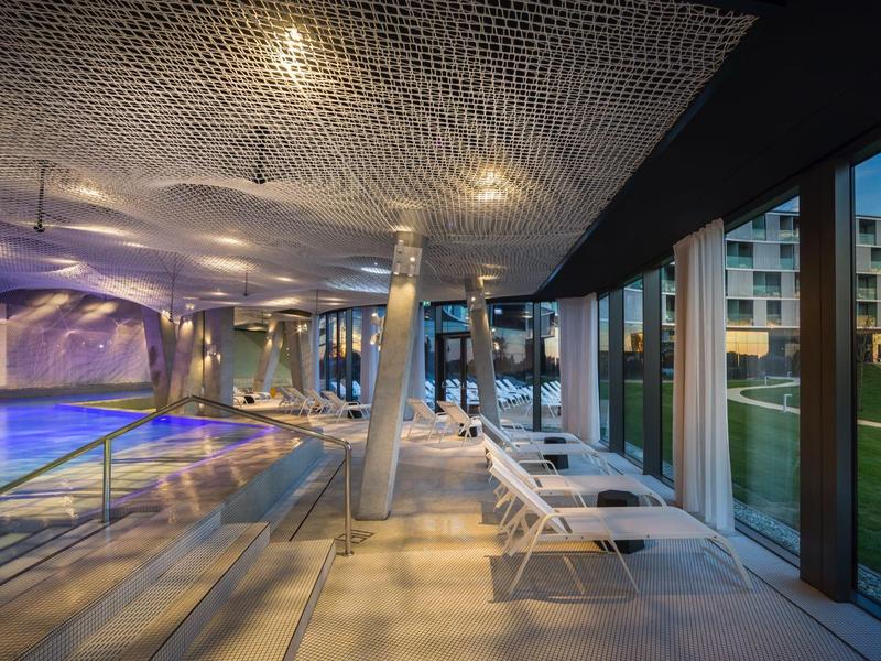 Moderner Hallenbadbereich mit Liegestühlen, Fensterfront und stimmungsvoller Beleuchtung.