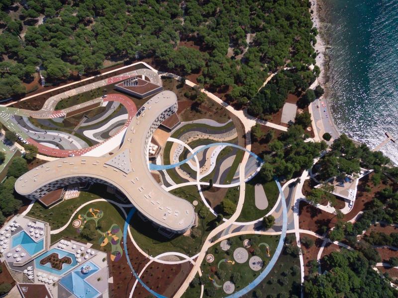 Luftaufnahme einer Küstenanlage mit geschwungenen Gebäuden, Pools, Wegen und umliegendem Wald.