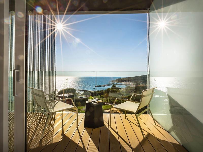Moderner Balkon mit Holzterrasse, zwei Stühlen und Blick aufs blaue Meer bei Sonnenschein.