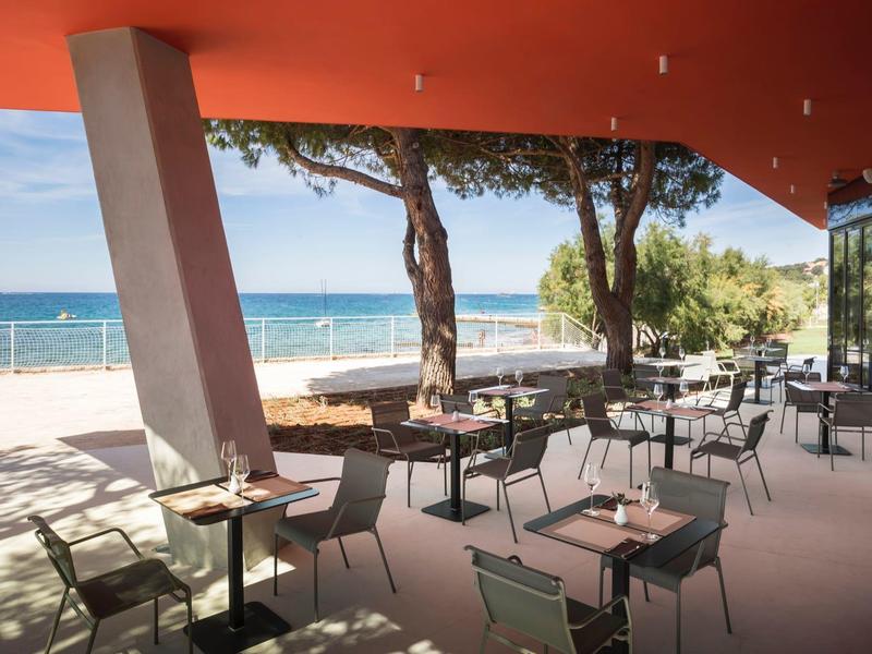 Terrassen-Café mit Tischen, Stühlen, Blick auf Meer, Sandstrand und Bäume bei sonnigem Himmel.