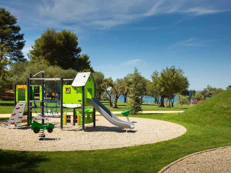Kinderspielplatz mit Rutsche, Klettergerüst und Wippen auf grüner Wiese unter blauem Himmel.