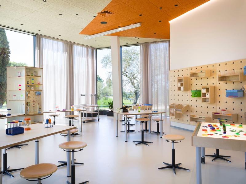 Heller, moderner Raum mit großen Fenstern, hohen Hockern und Tischen mit buntem Materialien.