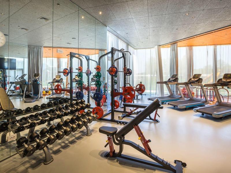 Moderne Fitnessstudio mit Hanteln, Laufbändern, Spiegelwand und großen Fenstern.