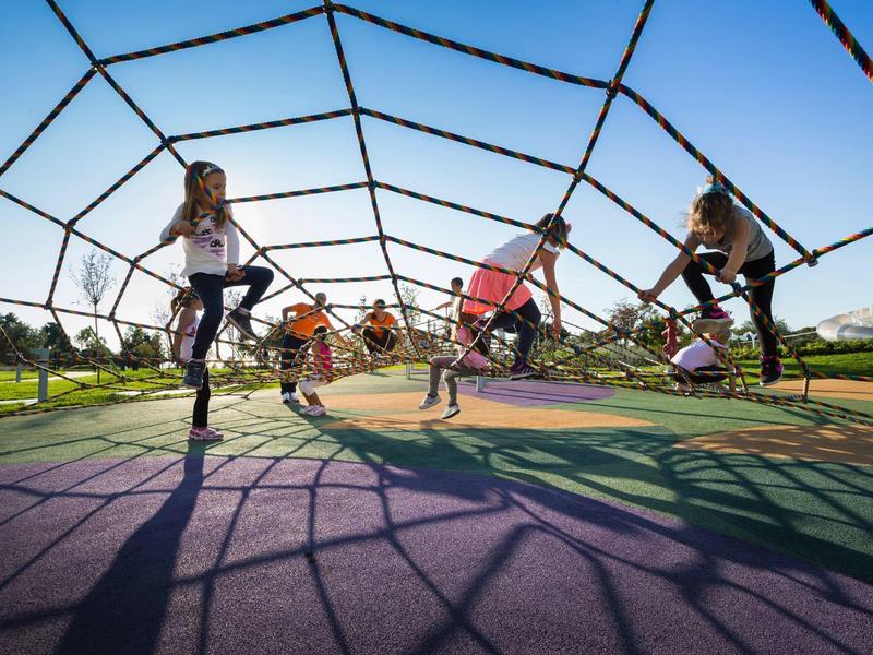 Kinder klettern auf einem großen Netzspielplatz im Freien bei blauem Himmel.