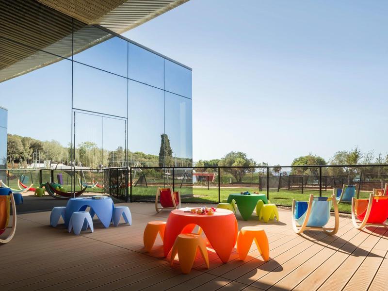 Moderner Außenbereich mit bunten Kunststoffstühlen und Holzboden, reflektierende Glasfassade und grüner Hintergrund.