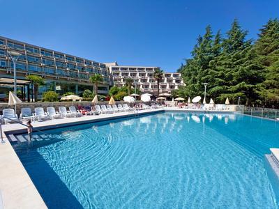 Grande piscina all'aperto con lettini davanti a un edificio hotel moderno e alberi verdi.