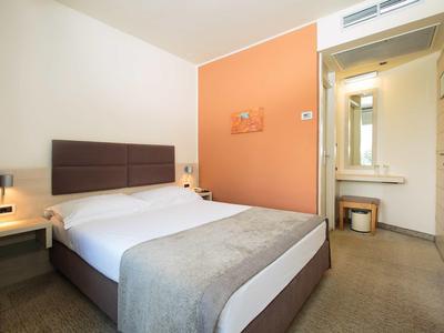 Moderne hotelkamer met tweepersoonsbed, oranje accentmuur en badkamer op de achtergrond.