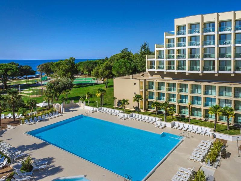 Groot hotel met zwembad, ligstoelen en uitzicht op zee onder een heldere hemel.