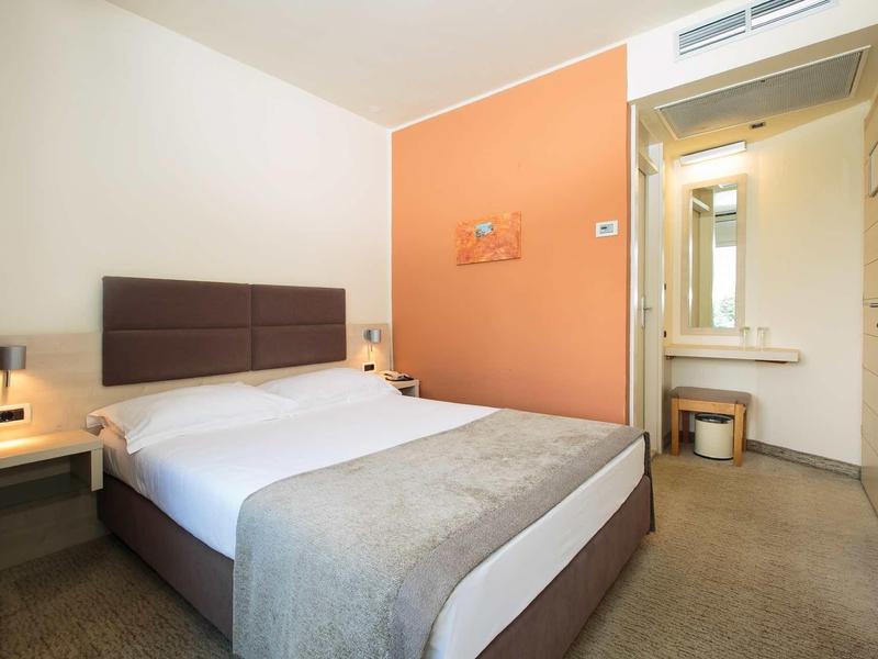 Moderne hotelkamer met tweepersoonsbed, oranje accentmuur en badkamer op de achtergrond.