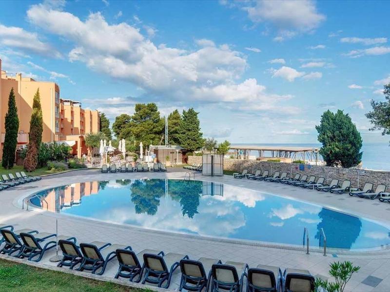 Grande piscina con sdraio davanti a un hotel su più piani sotto un cielo azzurro.