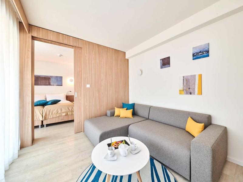 Soggiorno moderno con divano grigio e cuscini colorati accanto alla camera da letto aperta.