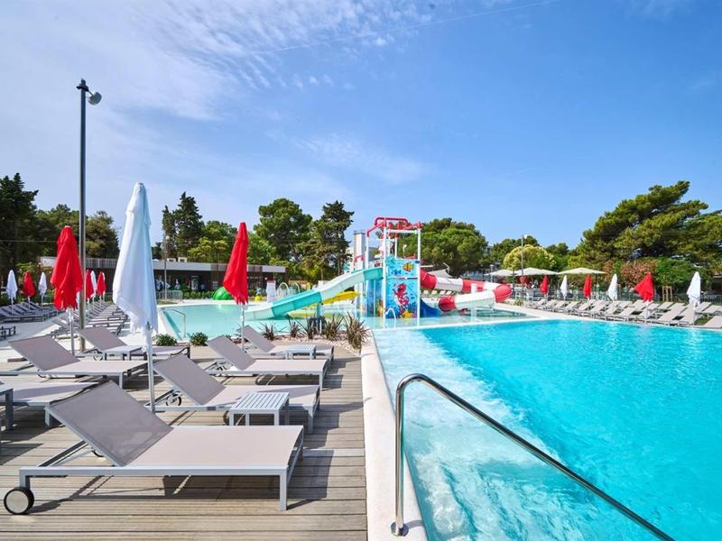 Piscina all'aperto luminosa con lettini, ombrelloni rossi e parco acquatico sullo sfondo.