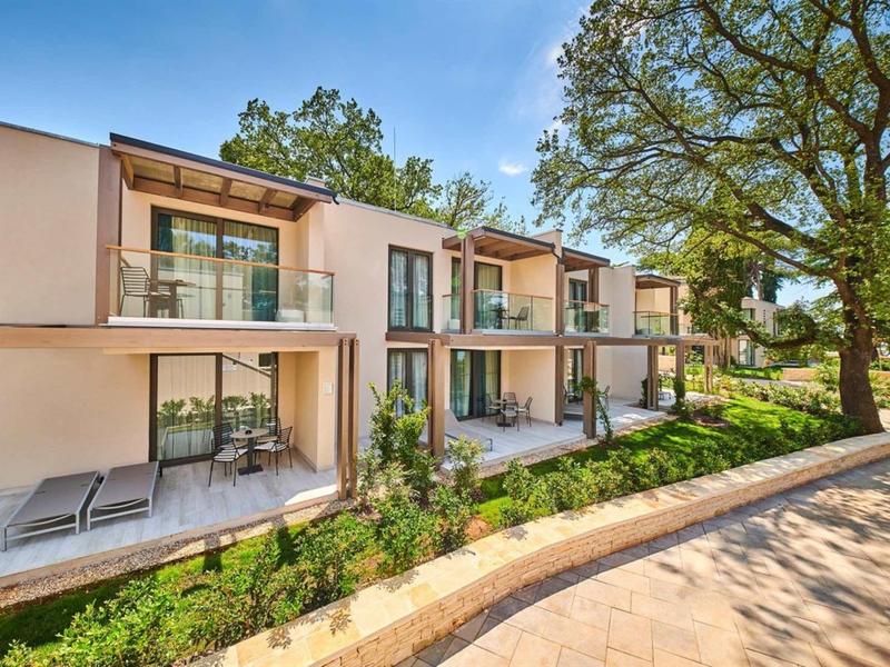 Complesso alberghiero moderno con terrazze e sdraio accanto a un sentiero e grandi alberi.