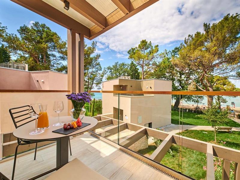 Balcone con tavolo, sedie e vista su giardino con alberi e edificio sotto cielo azzurro.