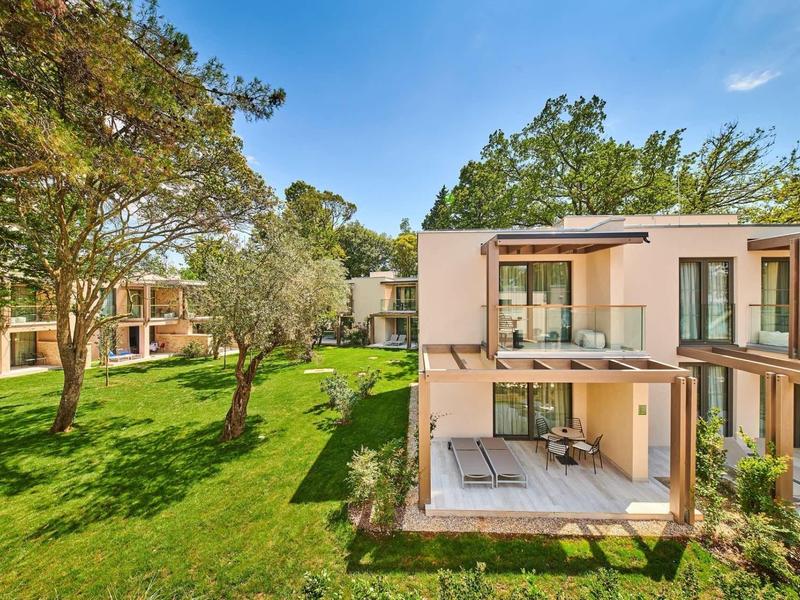 Bungalow moderno con terrazza e grandi finestre circondato da prato e alberi sotto un cielo sereno.