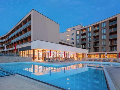 Modernes Hotel mit großen Glasfronten, mehreren Stockwerken und beleuchtetem Poolbereich bei Dämmerung.