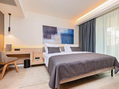 Modern eingerichtetes Hotelzimmer mit großem Bett, Schreibtisch und Balkonfenster mit Vorhängen.