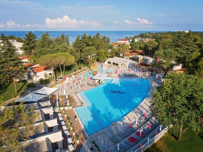 Ampia zona piscina con lettini e tavoli, circondata da alberi e vicino al mare.