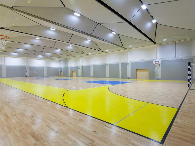 Grande salle de sport avec sol en bois et terrain de basket marqué en jaune sous un éclairage de plafond lumineux.