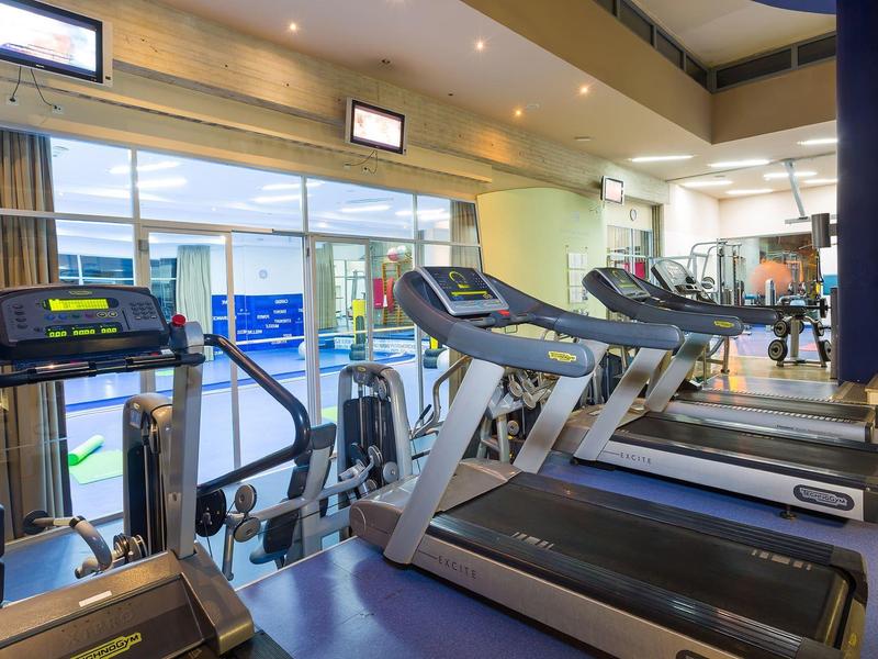 Salle de sport lumineuse avec tapis de course et appareils d'exercice, grandes fenêtres avec vue extérieure.