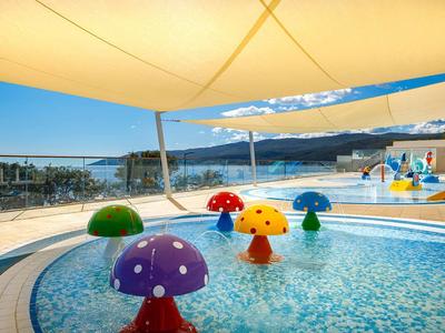 Children's pool with colorful mushroom water sprayers and shade sails overlooking the sea.