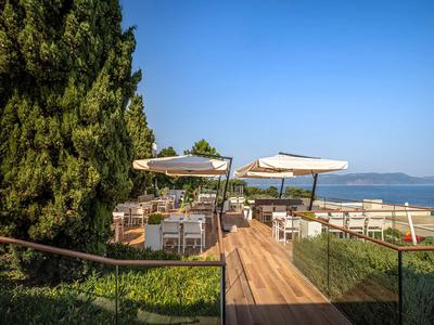 Houten pad leidt naar terras met tafels, stoelen en parasols, zee op de achtergrond.