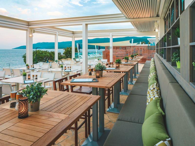 Restaurant terrace with wooden tables, cushions, and a clear sea view on a sunny day.