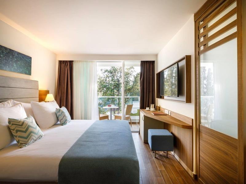 Modern hotel room with large bed, TV, wooden furniture, and balcony door overlooking greenery.