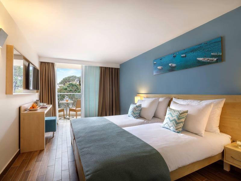 Modern hotel room with double bed, balcony, and view of trees and sky.