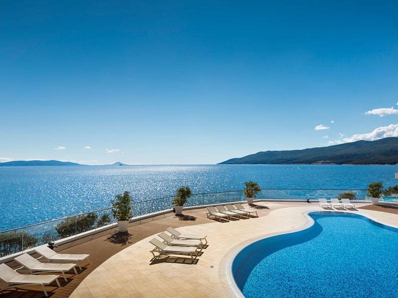 Pool with lounge chairs overlooking blue sea under clear sky.