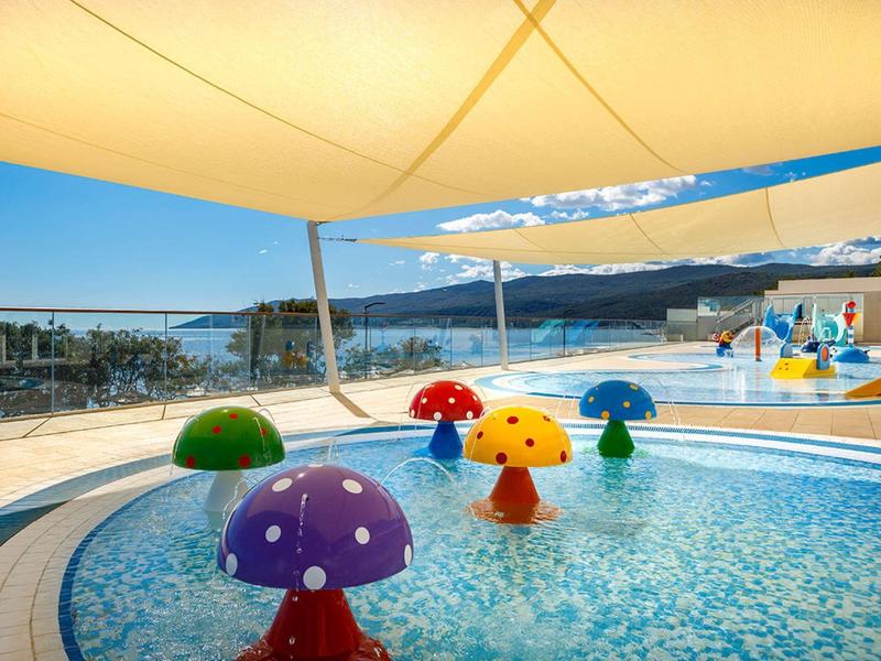 Children's pool with colorful mushroom water sprayers and shade sails overlooking the sea.