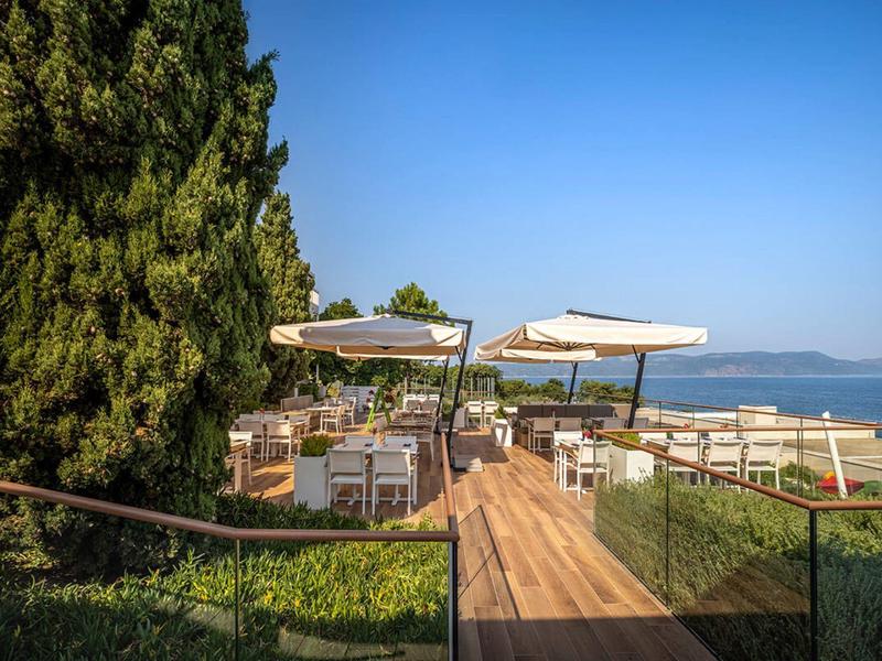 Terrace with tables, chairs, and umbrellas overlooking the sea and trees.