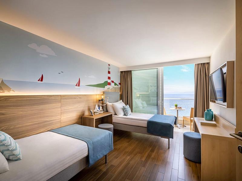 Modern hotel room with two single beds, wooden floor, and sea view through large window.